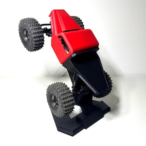 May include: A red and black remote control truck model with large, textured tires. The truck has a red body and a black chassis, and is displayed on a black stand. The tires are gray and have a rugged tread pattern.