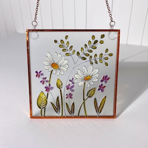 Puede incluir: Una ventana de vidrio tintado con marco de cobre que cuelga con un fondo blanco y un diseño floral con margaritas blancas con centros amarillos y flores moradas con hojas verdes.