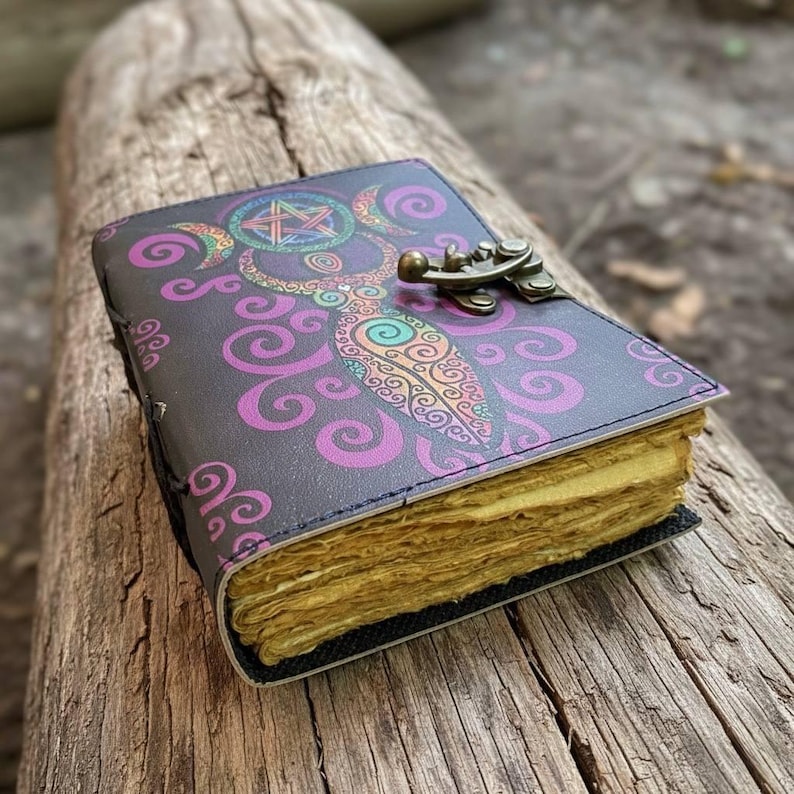 May include: A dark gray journal with a decorative cover featuring a colorful design of a crescent moon, star, and swirling patterns. The journal has a metal clasp and aged-looking pages. The journal is resting on a wooden log.