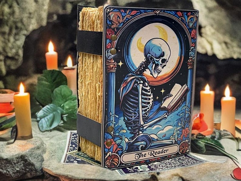 Peut inclure: Un livre rectangulaire avec une tranche en papier &eacute;pais et textur&eacute; brun clair, maintenu par des sangles noires. La couverture illustre un squelette lisant un livre sous un croissant de lune, avec le texte The Reader imprim&eacute; en bas.