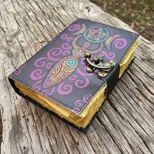 May include: A dark gray journal with a colorful, ornate design featuring a pentagram and crescent moons. The cover has a decorative pattern in purple, orange, and teal. It has a metal clasp and aged-looking pages.