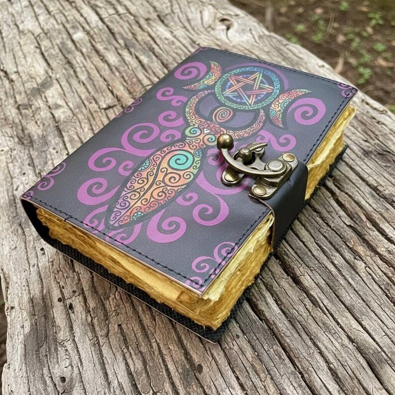 May include: A dark gray journal with a colorful, ornate design featuring a pentagram and crescent moons. The cover has a decorative pattern in purple, orange, and teal. It has a metal clasp and aged-looking pages.