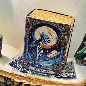 Peut inclure: Un livre rectangulaire intitul&eacute; The Reader avec une illustration de couverture repr&eacute;sentant un squelette lisant un livre sous un croissant de lune. Le livre pr&eacute;sente une palette de couleurs bleu fonc&eacute; et orange, des fermoirs en m&eacute;tal dor&eacute;.