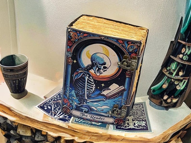 Peut inclure: Un livre rectangulaire intitul&eacute; The Reader avec une illustration de couverture repr&eacute;sentant un squelette lisant un livre sous un croissant de lune. Le livre pr&eacute;sente une palette de couleurs bleu fonc&eacute; et orange, des fermoirs en m&eacute;tal dor&eacute;.