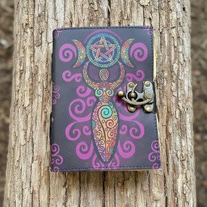 May include: A black journal with a colorful illustration of a goddess figure, crescent moons, and a pentagram. The cover features a decorative design in purple, orange, and green. It has a metal clasp closure.