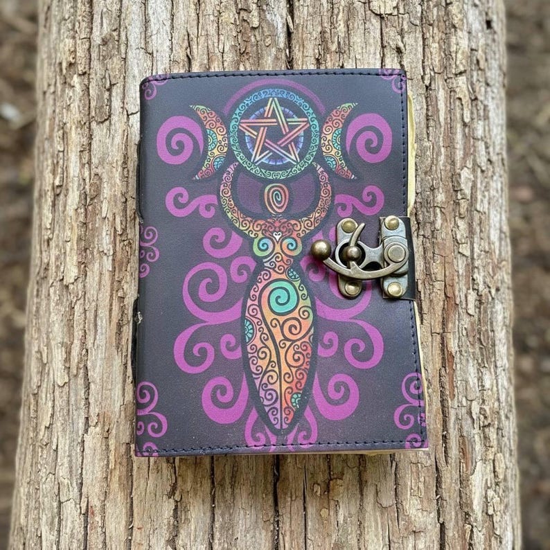 May include: A black journal with a colorful illustration of a goddess figure, crescent moons, and a pentagram. The cover features a decorative design in purple, orange, and green. It has a metal clasp closure.