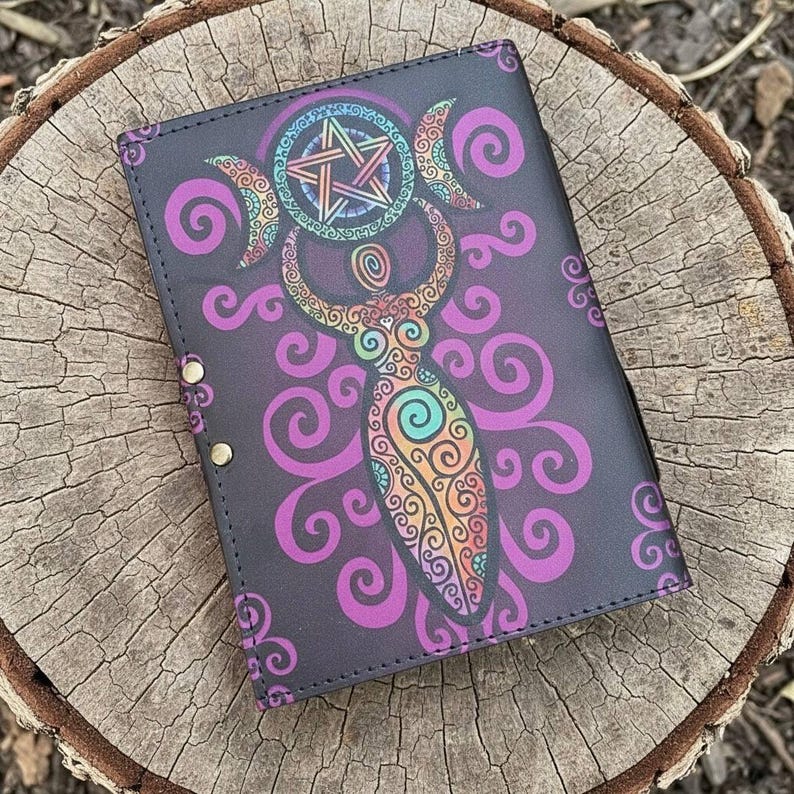 May include: A black journal cover featuring a colorful illustration of a goddess figure with a pentagram and crescent moons. The design includes purple swirls and intricate patterns. The journal is secured with brass-colored snaps.
