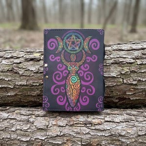 May include: A black journal with a colorful, intricate design featuring a pentagram, crescent moons, and a stylized figure. The design incorporates rainbow and purple hues. The journal is closed and has a snap closure.