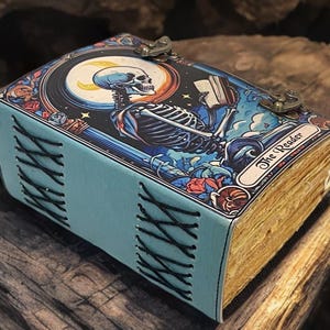 Peut inclure: Un journal rectangulaire avec une couverture bleu clair pr&eacute;sentant une illustration graphique d'un squelette lisant un livre sous un croissant de lune. La couverture est marqu&eacute;e The Reader et ferm&eacute;e par deux loquets m&eacute;talliques. Le dos est cousu en croix.
