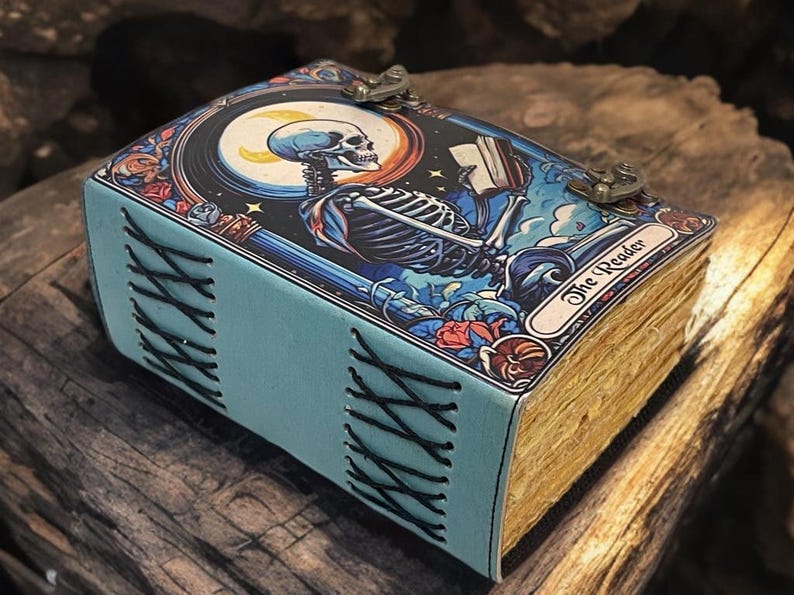 Peut inclure: Un journal rectangulaire avec une couverture bleu clair pr&eacute;sentant une illustration graphique d'un squelette lisant un livre sous un croissant de lune. La couverture est marqu&eacute;e The Reader et ferm&eacute;e par deux loquets m&eacute;talliques. Le dos est cousu en croix.