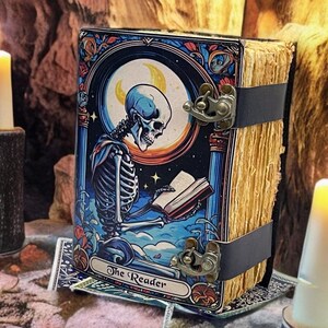 Peut inclure: Un livre rectangulaire intitul&eacute; The Reader pr&eacute;sentant une illustration color&eacute;e d'un squelette lisant un livre sous un croissant de lune. Le livre a une texture de pages vieillies et est fix&eacute; par deux fermoirs en m&eacute;tal fonc&eacute; sur le c&ocirc;t&eacute;.