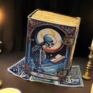 Peut inclure: Un livre rectangulaire avec une couverture en m&eacute;tal illustr&eacute;e d'un squelette lisant sous un croissant de lune. La couverture porte l'inscription The Reader. Le livre pr&eacute;sente des pages jaunies et est ferm&eacute; par deux loquets en laiton vieilli.