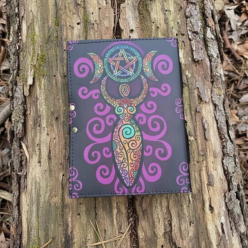 May include: A black journal with a colorful illustration of a goddess figure, crescent moons, and a pentagram. The design features purple, orange, teal, and yellow accents. The journal is closed with three silver snaps.