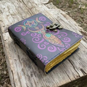 May include: A dark gray leather journal with a colorful illustration of a goddess figure and a pentagram. The cover features a brass clasp and is surrounded by purple swirls. The pages have a yellowed, aged appearance.