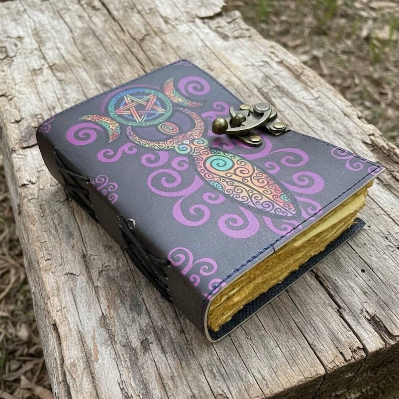 May include: A dark gray leather journal with a colorful illustration of a goddess figure and a pentagram. The cover features a brass clasp and is surrounded by purple swirls. The pages have a yellowed, aged appearance.