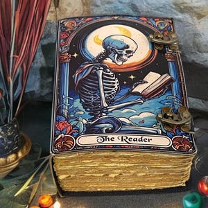 Peut inclure: Un journal rectangulaire &agrave; couverture rigide orn&eacute; d'une illustration color&eacute;e repr&eacute;sentant un squelette lisant un livre sous un croissant de lune. La couverture porte le texte The Reader et deux fermoirs en m&eacute;tal de couleur laiton.