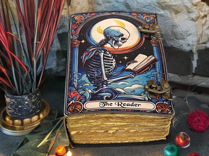 Peut inclure: Un journal rectangulaire &agrave; couverture rigide orn&eacute; d'une illustration color&eacute;e repr&eacute;sentant un squelette lisant un livre sous un croissant de lune. La couverture porte le texte The Reader et deux fermoirs en m&eacute;tal de couleur laiton.