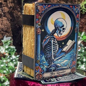 Peut inclure: Un livre &agrave; couverture rigide intitul&eacute; The Reader avec une illustration color&eacute;e d'un squelette lisant un livre sous un croissant de lune. Le livre a des pages &eacute;paisses, textur&eacute;es et brun clair maintenues par deux sangles noires et des rivets.