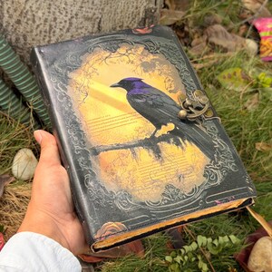 Puede incluir: Un diario rectangular con cubierta negra que presenta un borde en relieve ornamentado y un gr&aacute;fico central de un cuervo negro posado en una rama sobre un fondo amarillo. El diario incluye un cierre de pestillo met&aacute;lico en el lado derecho.
