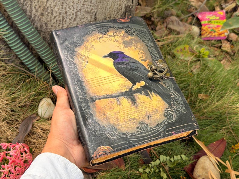 Puede incluir: Un diario rectangular con cubierta negra que presenta un borde en relieve ornamentado y un gr&aacute;fico central de un cuervo negro posado en una rama sobre un fondo amarillo. El diario incluye un cierre de pestillo met&aacute;lico en el lado derecho.
