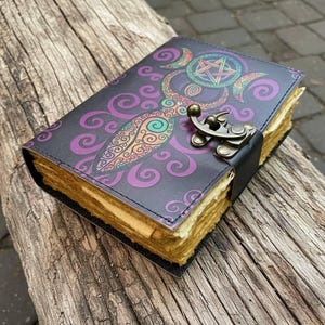May include: A leather-bound journal with a decorative cover featuring a purple and teal design with a pentagram and crescent moons. The journal has a metal clasp and aged paper pages. The journal is resting on a weathered wooden surface.