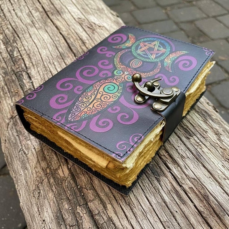 May include: A leather-bound journal with a decorative cover featuring a purple and teal design with a pentagram and crescent moons. The journal has a metal clasp and aged paper pages. The journal is resting on a weathered wooden surface.