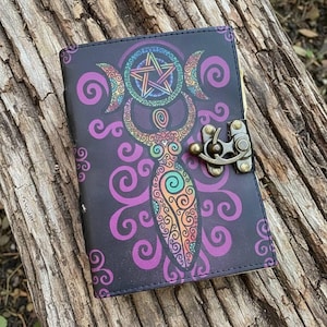 May include: A black journal with a colorful illustration of a goddess, crescent moons, and a pentagram. The design features purple, orange, green, and blue accents. The journal has a metal clasp and is closed.