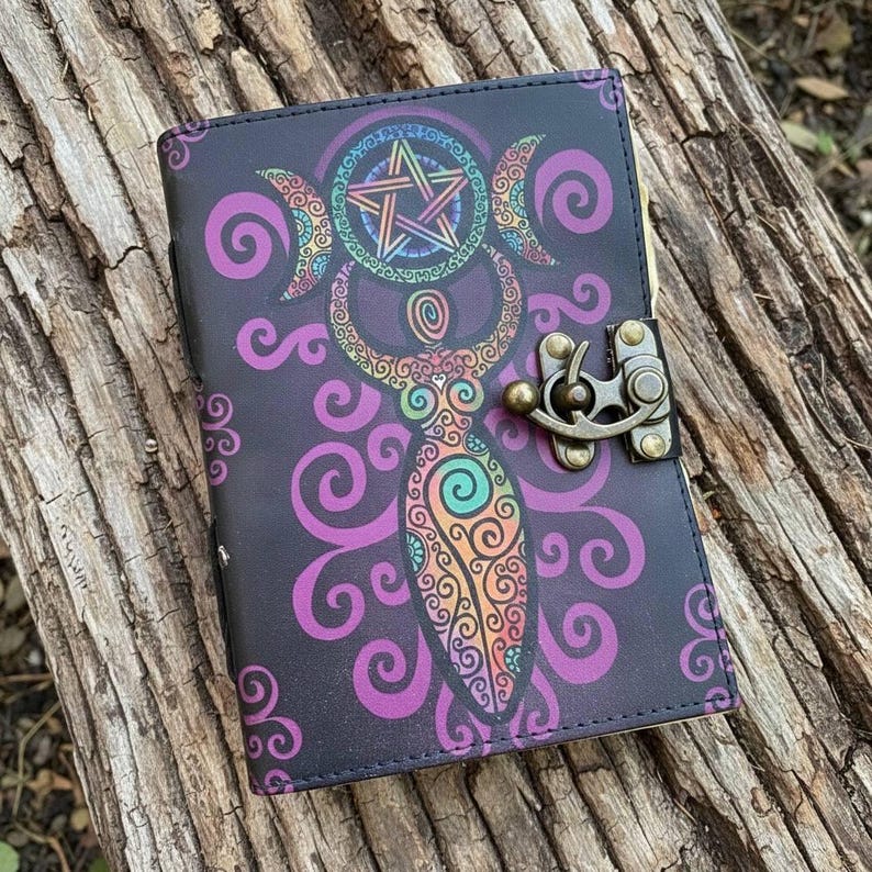 May include: A black journal with a colorful illustration of a goddess, crescent moons, and a pentagram. The design features purple, orange, green, and blue accents. The journal has a metal clasp and is closed.