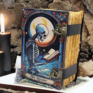 Peut inclure: Un livre reli&eacute; intitul&eacute; The Reader avec une illustration color&eacute;e d'un squelette lisant un livre sous un croissant de lune. Le livre a des pages vieillies et est maintenu par deux sangles noires avec des fermoirs en m&eacute;tal laitonn&eacute;.