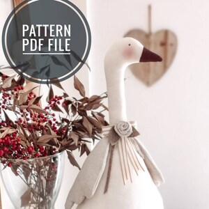Può includere: Un'oca di stoffa bianca con un becco marrone e un blush rosa sulle guance. L'oca indossa un fiocco marrone e beige con una rosa bianca. L'oca è seduta su una superficie bianca. Il testo "PATTERN PDF FILE" è in un cerchio nell'angolo in alto a sinistra dell'immagine.