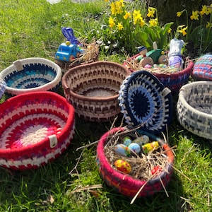 Könnte beinhalten: Eine Sammlung handgefertigter gewebter Körbe in verschiedenen Farben, darunter Rot, Blau und Naturtöne. Einige Körbe enthalten dekorative Gegenstände wie Spielzeugautos und bemalte Eier, die auf Gras platziert sind. Die Körbe sind rund und haben eine strukturierte Oberfläche.