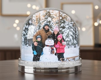 Puede incluir: Un globo de nieve con un retrato familiar y un muñeco de nieve en un paisaje de bosque nevado. La familia lleva ropa de invierno. El globo se apoya en una superficie de madera oscura con una base plateada. Luces decorativas en el fondo.
