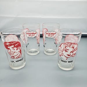 May include: A white ceramic mug with a black handle and the text "But First, Coffee" in a bold, black font. The mug is filled with a dark liquid, likely coffee, and sits on a light-colored surface.