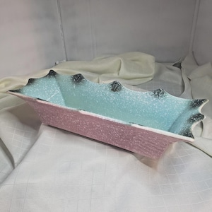 May include: A rectangular ceramic dish with a pink exterior and a light blue interior. The rim has a scalloped edge with black accents. The dish is likely for decorative or serving purposes.