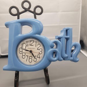 May include: A light blue "Bath" clock with a round clock face. The clock face has black numbers and hands, and the words "New Haven Quartz" are printed on the face. The clock is supported by a black metal stand.
