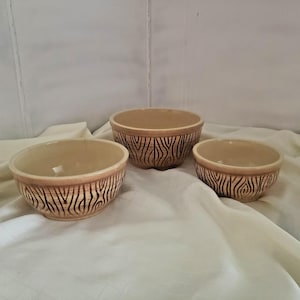 May include: Three beige ceramic bowls with a textured, wood-grain-like pattern around the rims. The bowls vary in size, with a smooth interior and a neutral color palette. They are arranged on a white cloth.