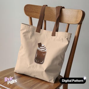 Puede incluir: Bolso tote beige con asas marrones, con un gráfico pixelado de una bebida de chocolate con crema batida y una galleta. Las palabras "Digital Pattern" están impresas en la parte inferior derecha.