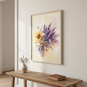 May include: Watercolour print of a bouquet featuring a sunflower, lavender, and daisies, tied with a brown ribbon. The artwork is framed in a light wood frame and hangs on a cream-coloured wall. Bees are scattered around the flowers.