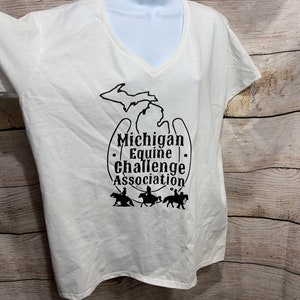 Könnte beinhalten: Weißes T-Shirt mit V-Ausschnitt mit einer schwarzen Silhouette des Bundesstaates Michigan und dem Text "Michigan Equine Challenge Association" mit vier Reitern auf Pferden.