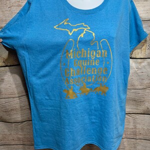 Könnte beinhalten: Hellblaues T-Shirt mit goldener Grafik, die den Umriss von Michigan und den Text "Michigan Equine Challenge Association" zeigt. Das Shirt hat einen Rundhalsausschnitt und kurze Ärmel.