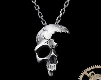 Könnte beinhalten: Silberfarbener Totenkopf-Anhänger mit Kette. Der Schädel hat ein Riss-Design auf der Oberseite und detaillierte Gesichtszüge. Der Anhänger hängt vor einem schwarzen Hintergrund.