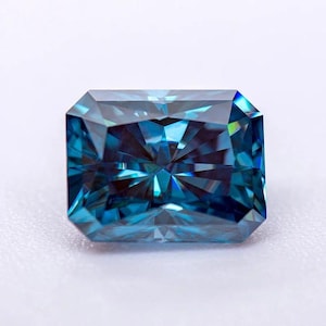 May include: A rectangular, faceted gemstone with a deep teal color. The gem has a brilliant cut, reflecting light to create a sparkling effect. The gemstone is set against a white background, highlighting its color and clarity.