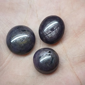 Peut inclure: Trois pierres précieuses polies, de couleur violet foncé, avec un effet étoilé. Les pierres sont ovales et rondes, avec une surface réfléchissante. Les gemmes sont posées sur un fond neutre, mettant en valeur leur couleur et leur texture.