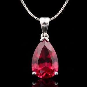 Peut inclure: Un collier en argent avec un pendentif en forme de goutte d'eau, facetté, en pierre rouge. Le pendentif est suspendu à une chaîne délicate sur fond noir. La pierre précieuse est taillée pour refléter la lumière.