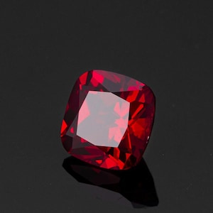 May include: A cushion-cut gemstone in a rich, deep red colour. The gem's faceted surface reflects light, creating a bright display. The gemstone is set against a dark background, accentuating its colour and shape.