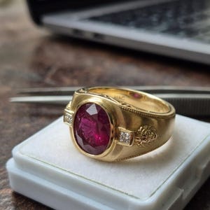May include: A gold ring featuring a large, oval-cut ruby gemstone. The ring has a detailed design with small diamonds on each side of the ruby and a lion's head on the band. The ring is set on a white surface.