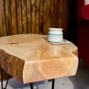 Puede incluir: Una mesa auxiliar de madera natural con borde vivo. La mesa tiene un veteado claro y está sostenida por patas de horquilla de metal negro. Un pequeño tarro blanco con rayas azules está sobre la mesa.