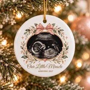 May include: A round white ceramic ornament featuring a central oval ultrasound image of a fetus, framed by a floral wreath and a pink bow. The text reads "Our Little Miracle" in script and "ARRIVING 2027" in block letters, with a gold hanging cord.
