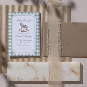 May include: A baby shower invitation with a green gingham border and a rocking horse illustration. The invitation includes the text "Baby Shower" and event details. A brown envelope and decorative elements complete the scene.