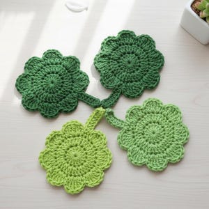 May include: Four handmade crochet coasters in shades of green, shaped like leaves. They are arranged on a light-coloured wooden surface. A small succulent plant in a white pot is visible in the background.